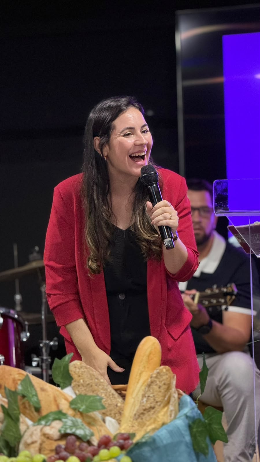 Mulher de casaco vermelho a sorrir ao microfone, atrás de um cesto de pães e uvas